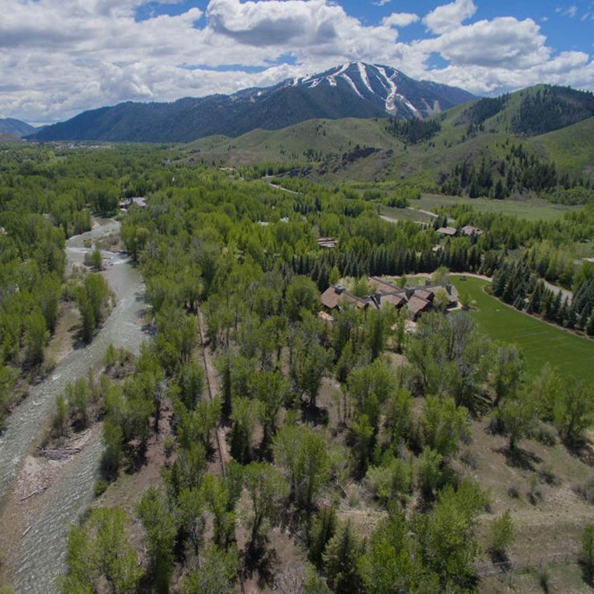Aerial view of a lush, green valley with a river winding through, surrounded by mountains and dotted with homes and trees under a partly cloudy sky.