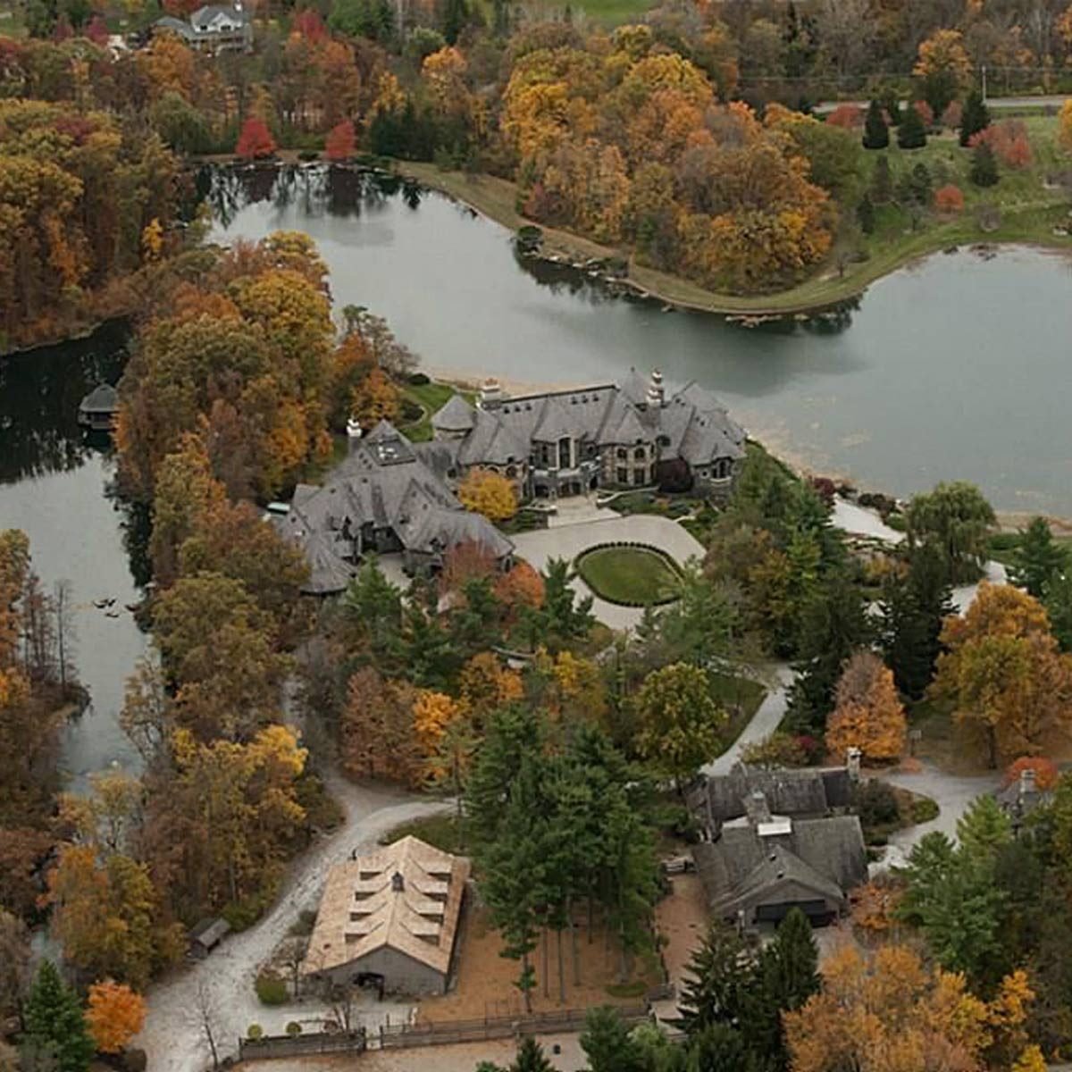 A large mansion sits near a serene lake, surrounded by colorful autumn trees, while winding paths and additional structures are visible nearby.