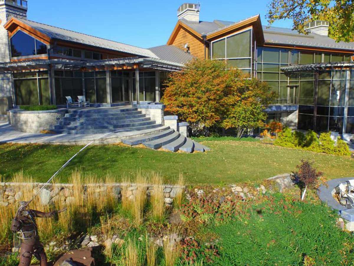 A modern, multi-level home with large windows overlooks a landscaped yard, featuring steps leading to a figure casting a fishing line near water.