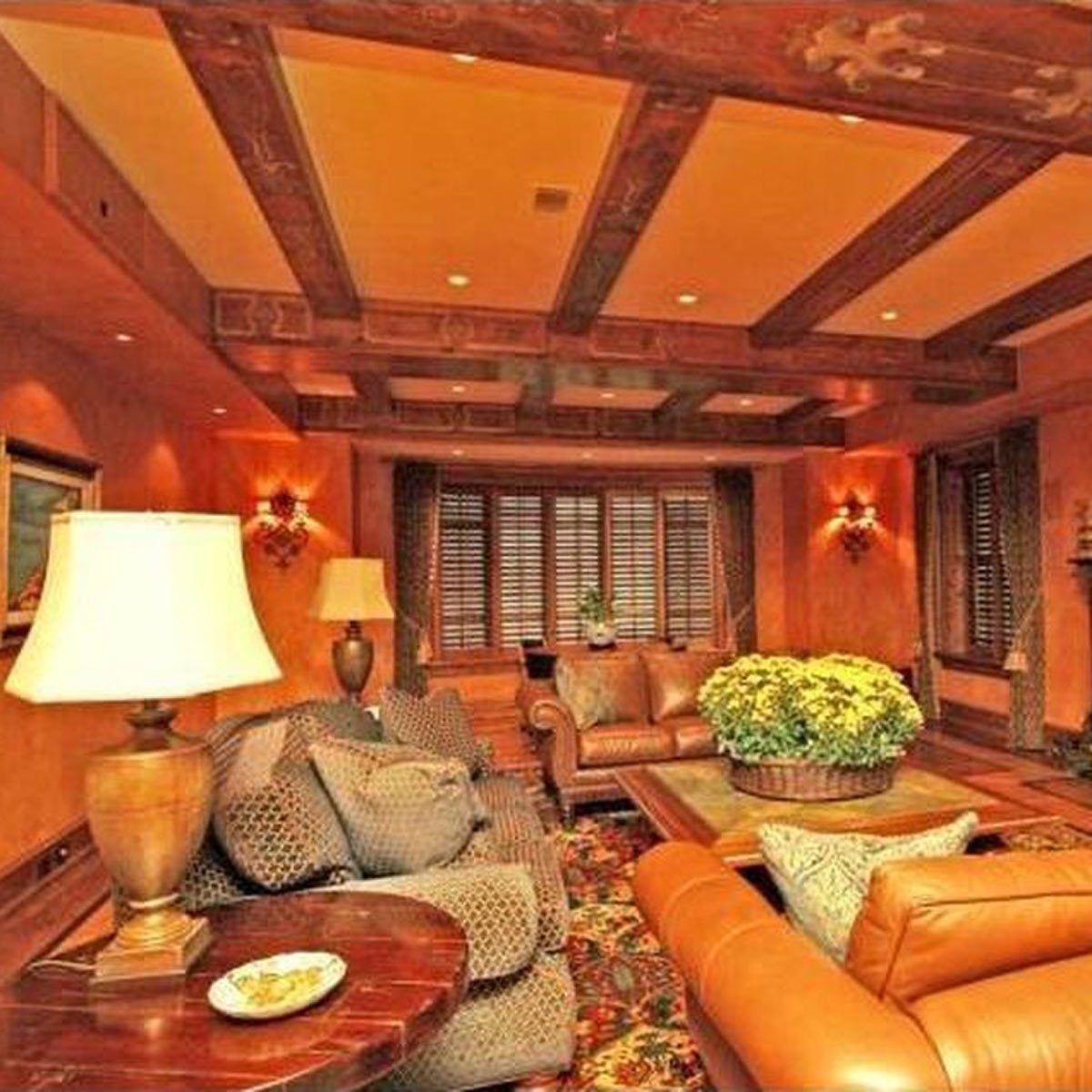 A cozy living room features plush sofas and a coffee table, illuminated by lamps, surrounded by warm-toned walls and wooden beams. A floral centerpiece decorates the table.