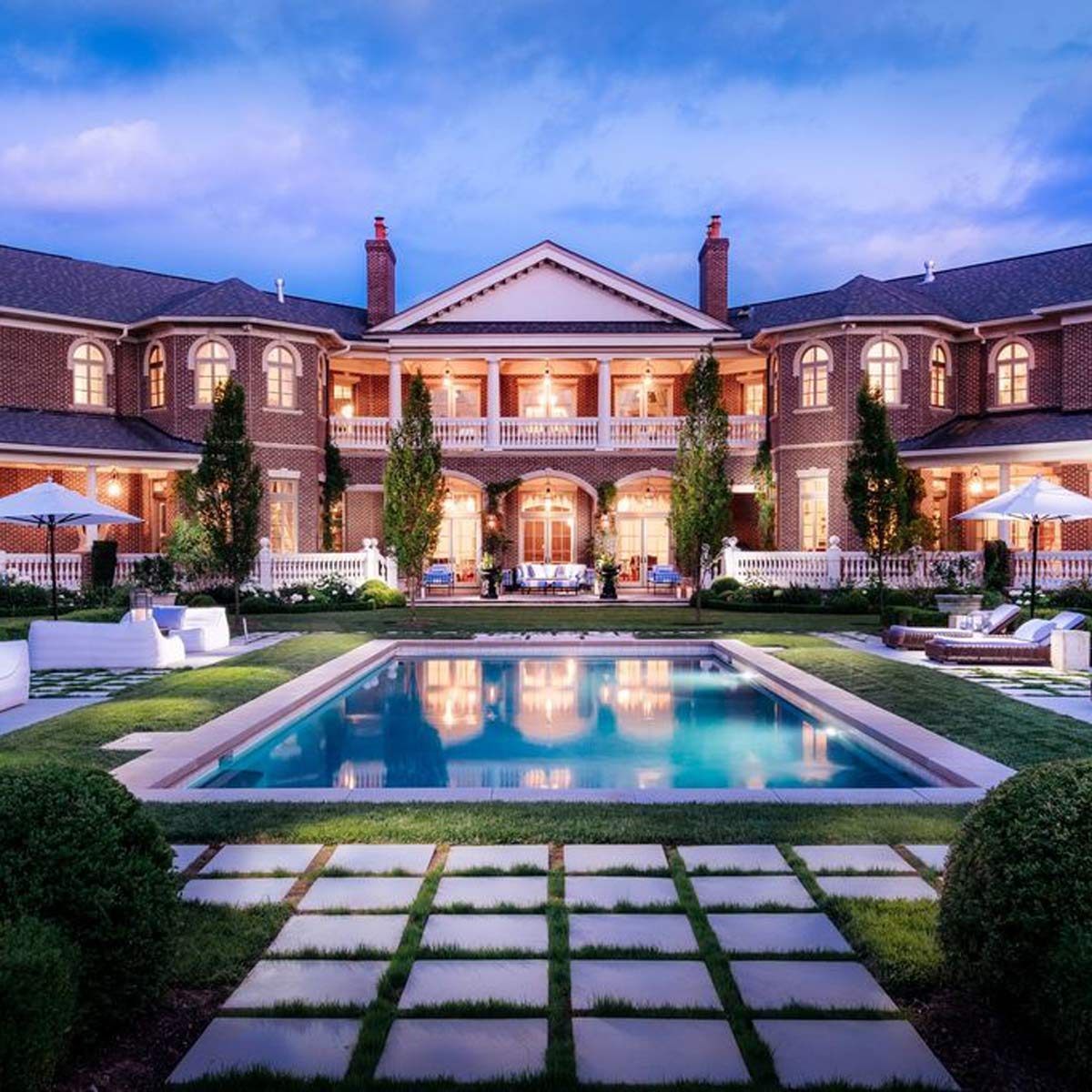 A luxurious mansion stands elegantly by a swimming pool. Soft lighting glows from large windows, surrounded by landscaped gardens and lounge areas under umbrellas.