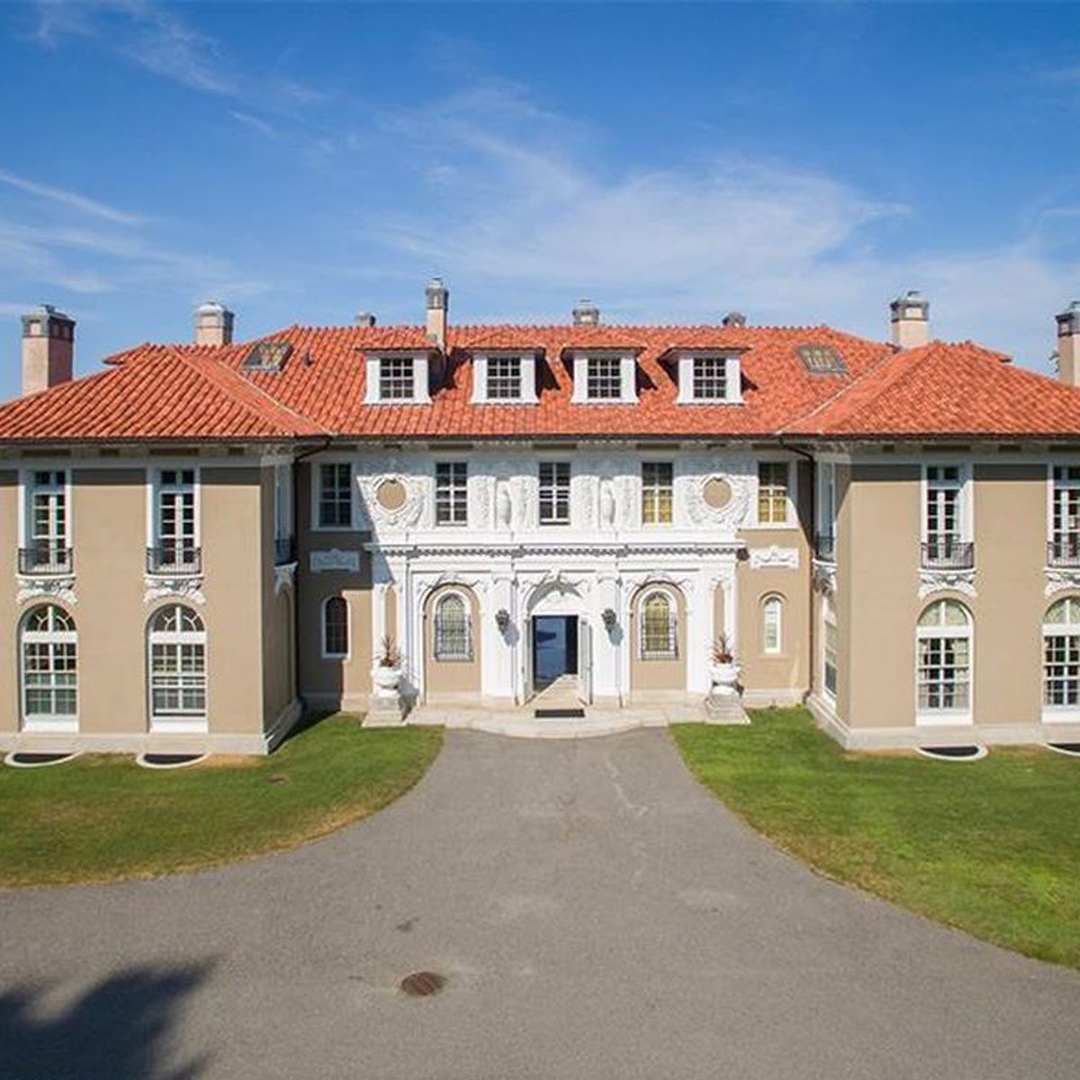A large, elegant mansion with a decorative fa&ccedil;ade sits on a manicured lawn, framed by blue skies and a gravel driveway leading to its entrance.