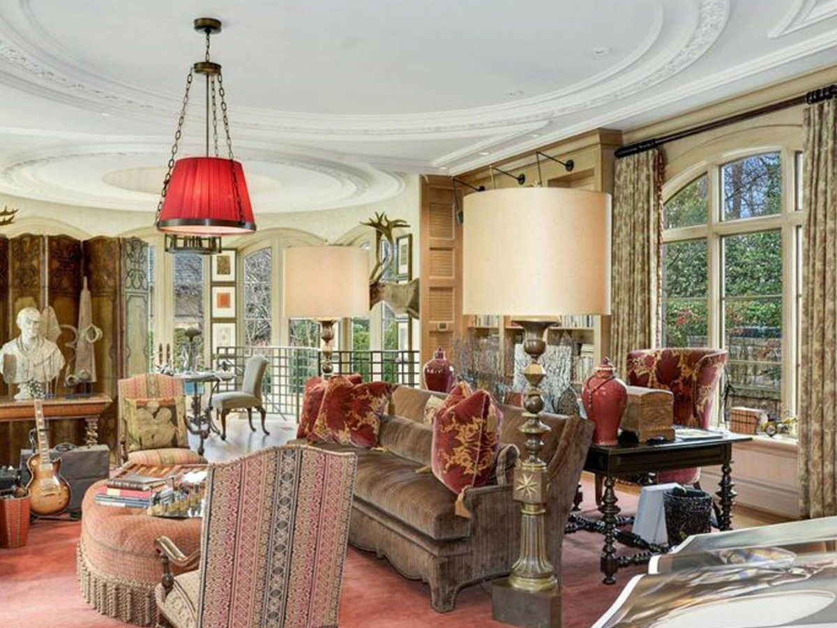 A warmly decorated living room features a plush sofa, armchairs, and unique lamps, illuminated by natural light from large windows, creating an inviting atmosphere.