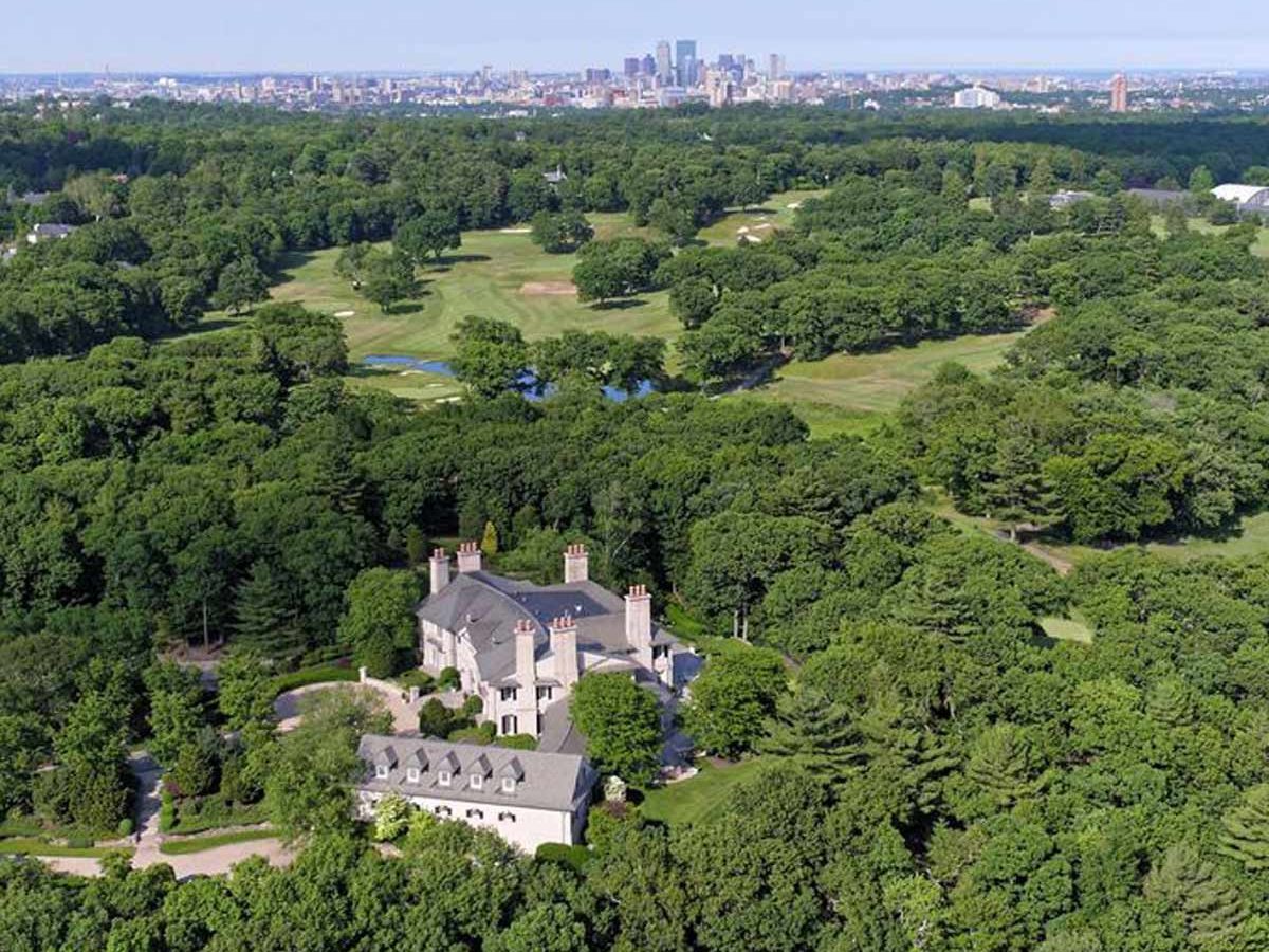 A large, elegant mansion is situated among dense greenery, while a golf course stretches out nearby, with a city skyline visible in the distance.
