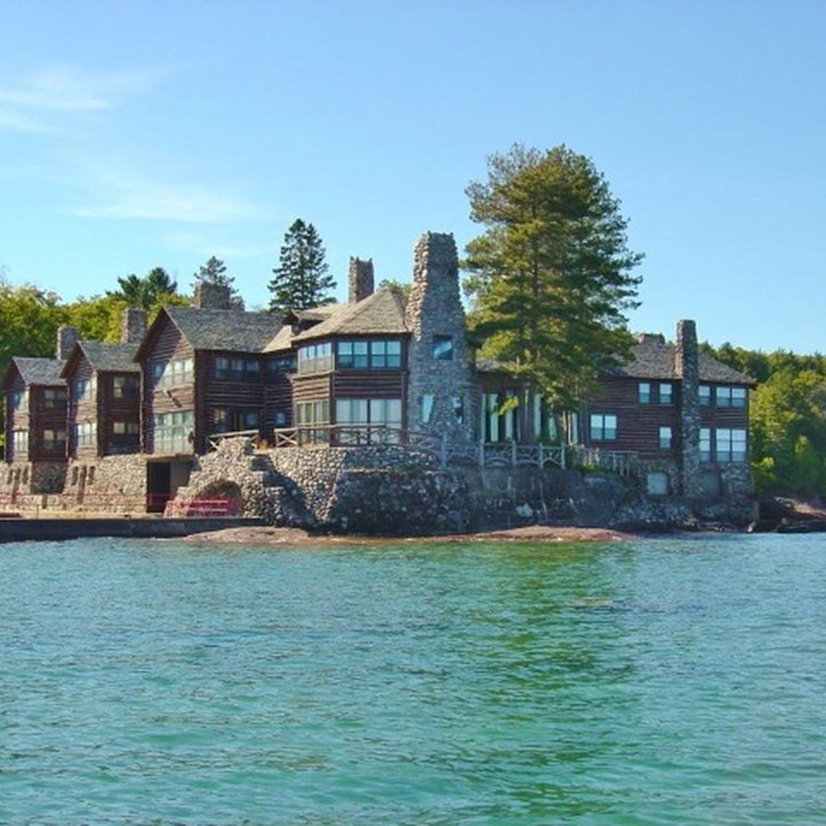A large stone and wood house stands on rocky shore, surrounded by trees, with clear water in the foreground and a blue sky above.