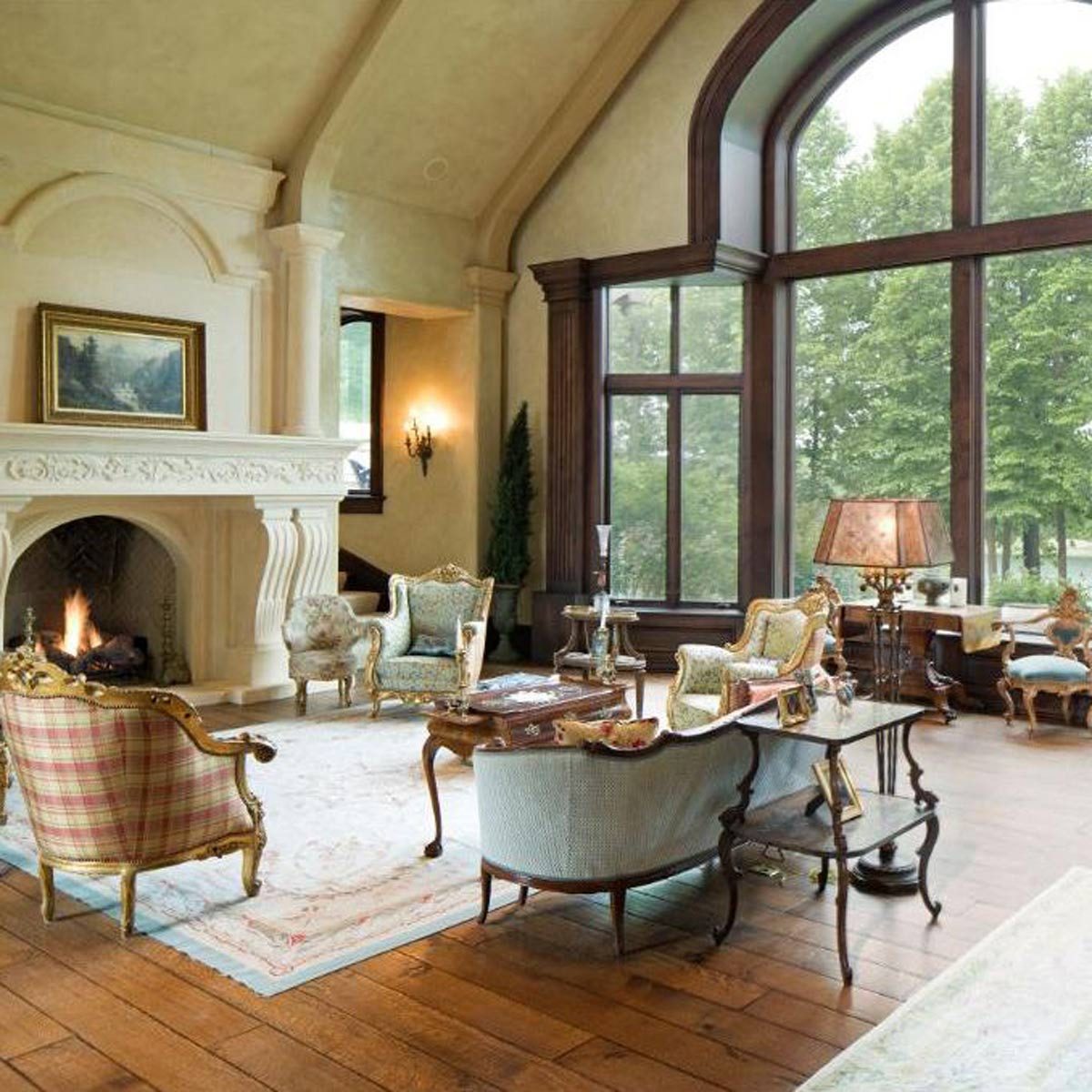 Elegant armchairs surround a wooden coffee table, as a fire burns in a grand fireplace, illuminated by natural light from tall windows.