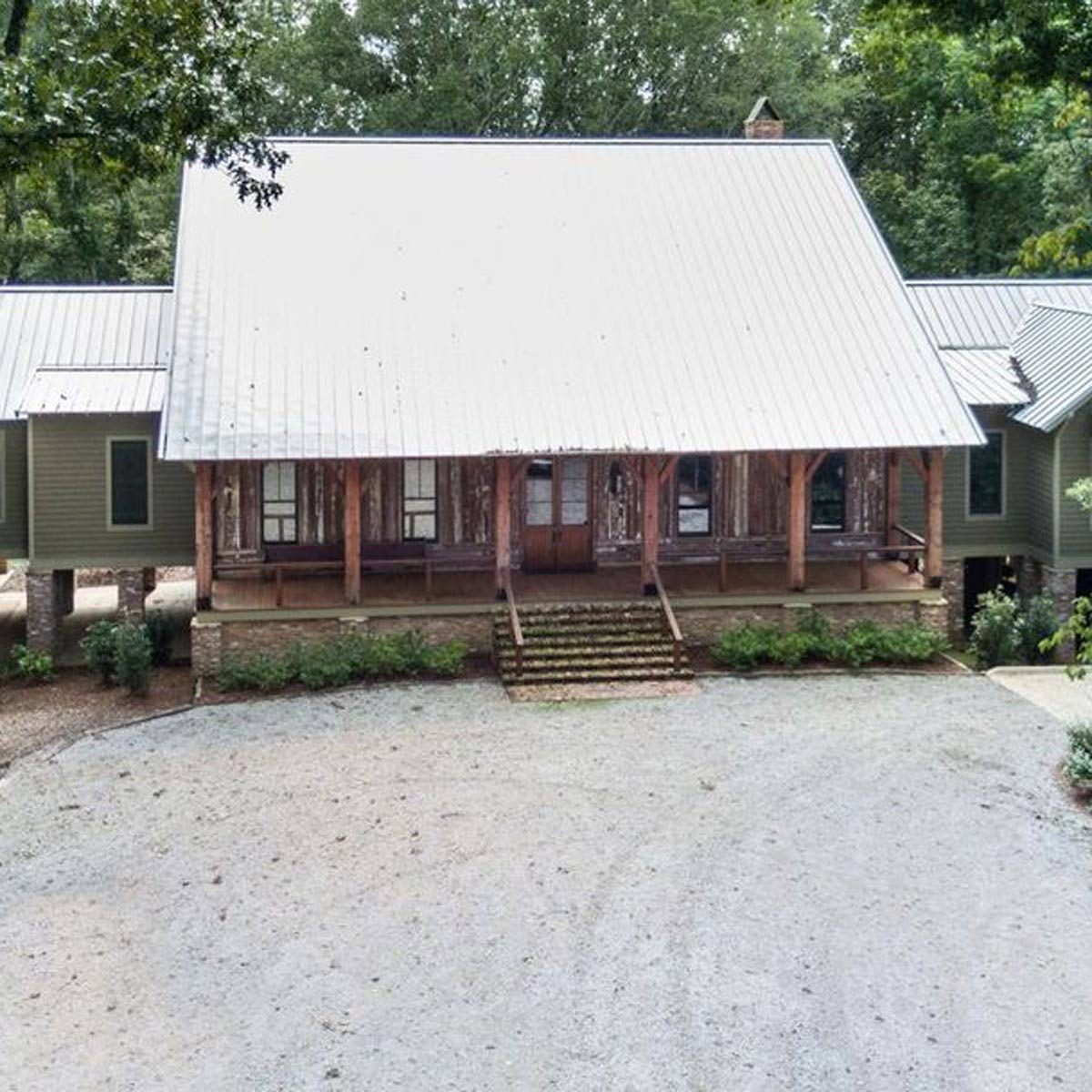 A large, rustic house with a metal roof stands on a gravel driveway, surrounded by greenery and trees, showcasing a welcoming entrance with steps.