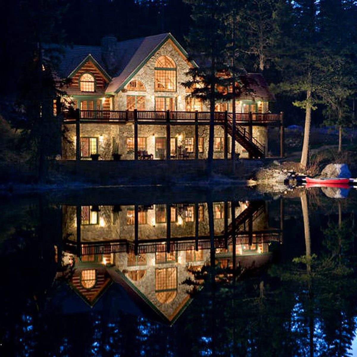 A large, illuminated stone house reflects on a calm lake at night, surrounded by dark trees and a red kayak nearby.