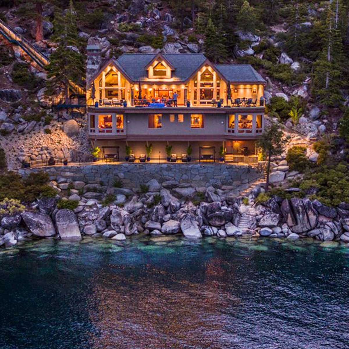 A large, illuminated lakeside house sits on rocky terrain, with people visible on the balcony, surrounded by trees and a calm waterway below.