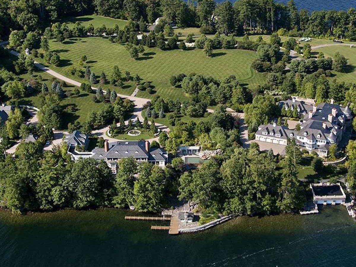 Aerial view of luxurious properties by a lake, surrounded by manicured green lawns and trees, with a dock extending into the water.