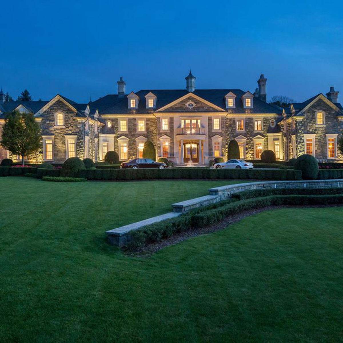A grand, illuminated mansion stands prominently in the evening, surrounded by well-manicured lawns and neatly trimmed hedges, with parked cars in the driveway.