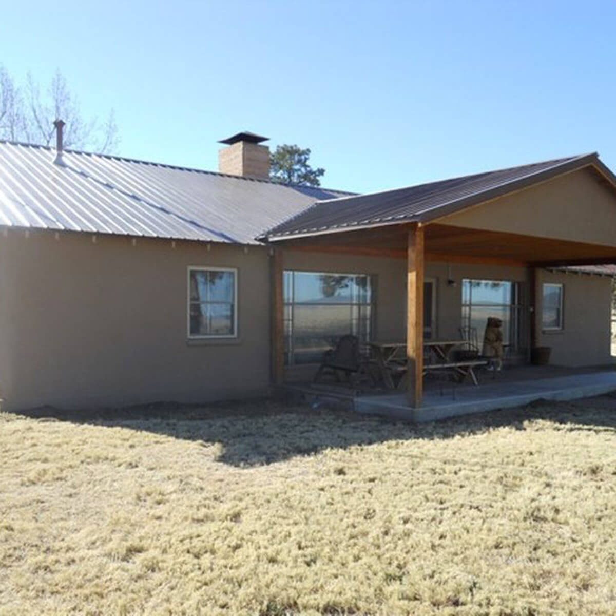 A single-story house with a metal roof features a covered porch and large windows. It sits in a grassy area under a clear blue sky.