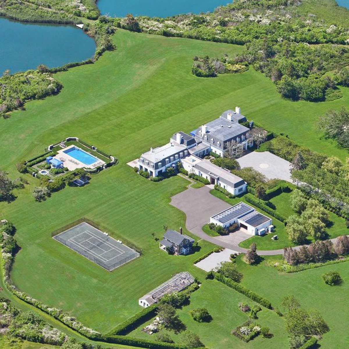 A large estate features a grand house, swimming pool, tennis court, and manicured lawns, surrounded by water and greenery in a spacious open landscape.