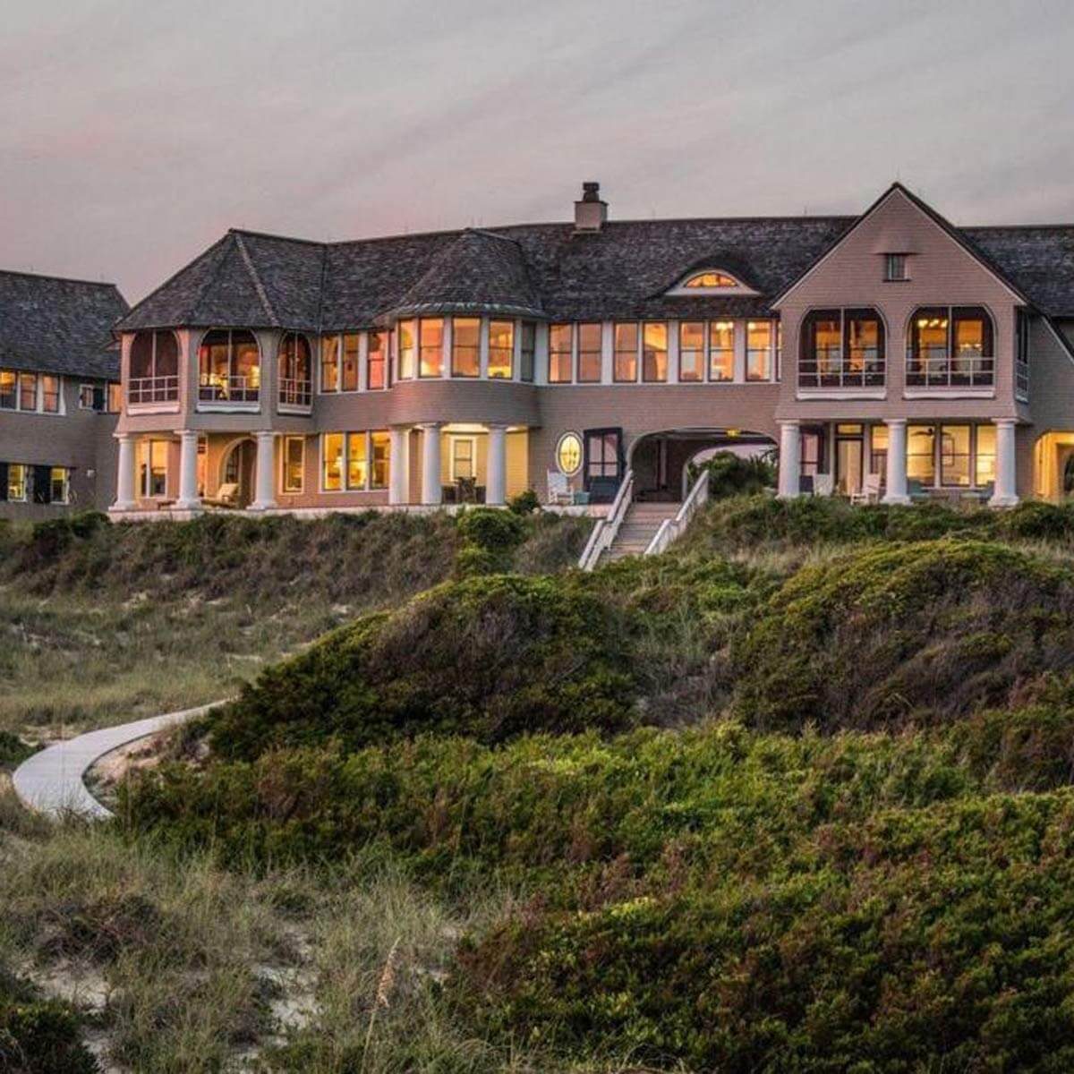 A large, elegant seaside house stands on a grassy hill, illuminated at dusk, with multiple windows and architectural detailing, surrounded by lush vegetation.