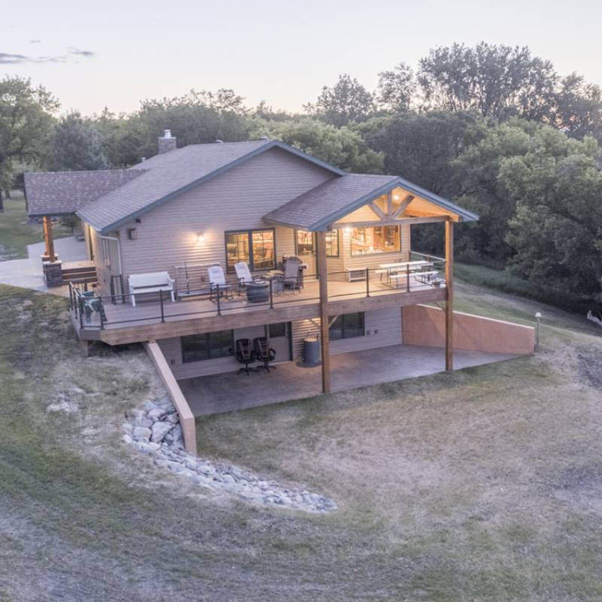 A modern house with a wooden deck overlooks a grassy hillside, surrounded by trees, illuminated by evening light, creating a cozy atmosphere.