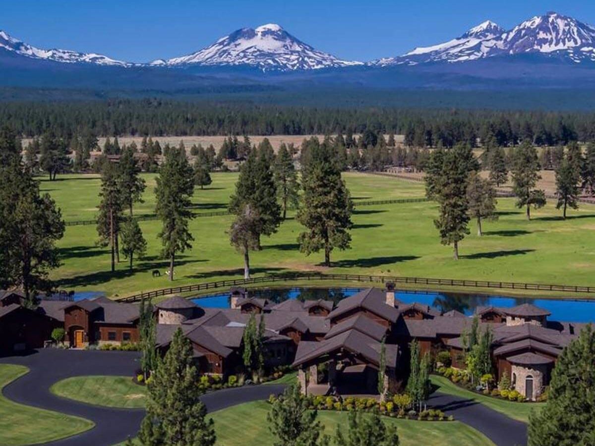 A luxurious collection of homes surrounds a circular driveway, bordered by trees and a pond, with majestic snow-capped mountains in the background under a clear blue sky.