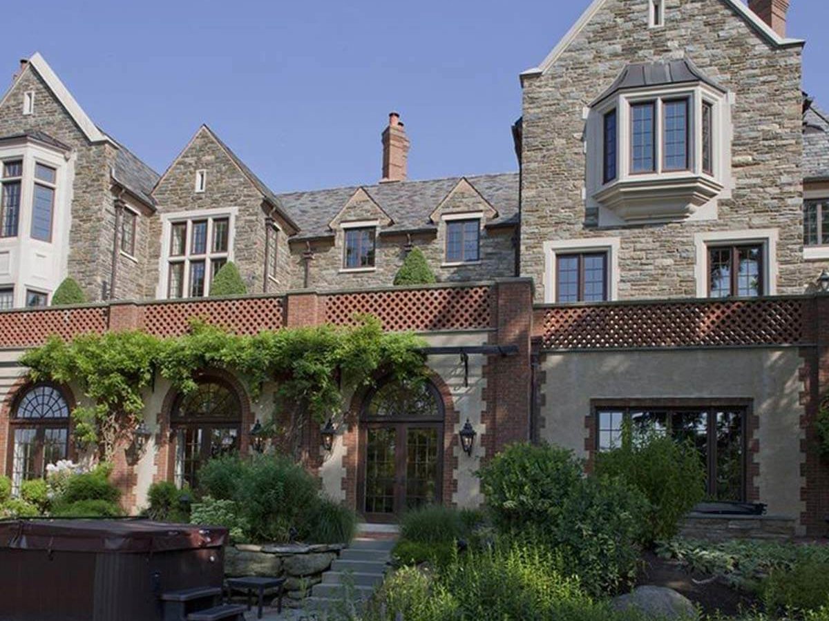 A large stone house features multiple windows and a lush garden. A hot tub sits nearby, surrounded by greenery under a clear blue sky.