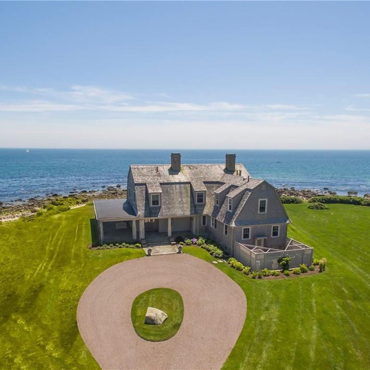 A large coastal house stands with a circular gravel driveway, overlooking the ocean, surrounded by green grass and rocky shoreline under a clear blue sky.