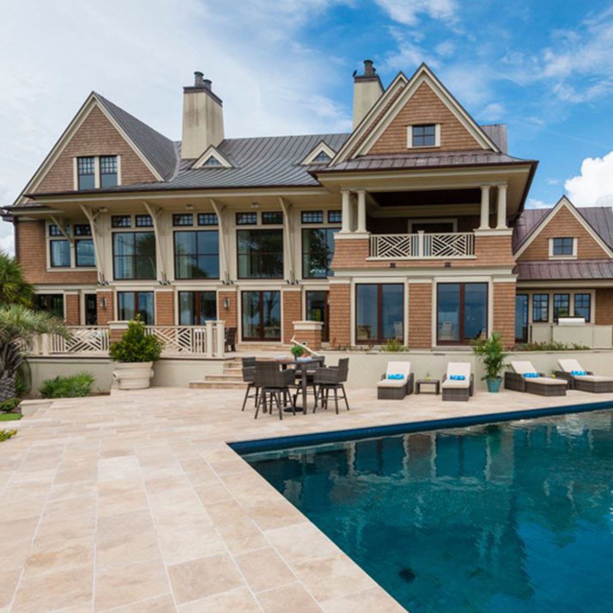 A large, elegant house with multiple windows overlooks a pool and patio area, featuring outdoor furniture and lush greenery under a cloudy blue sky.