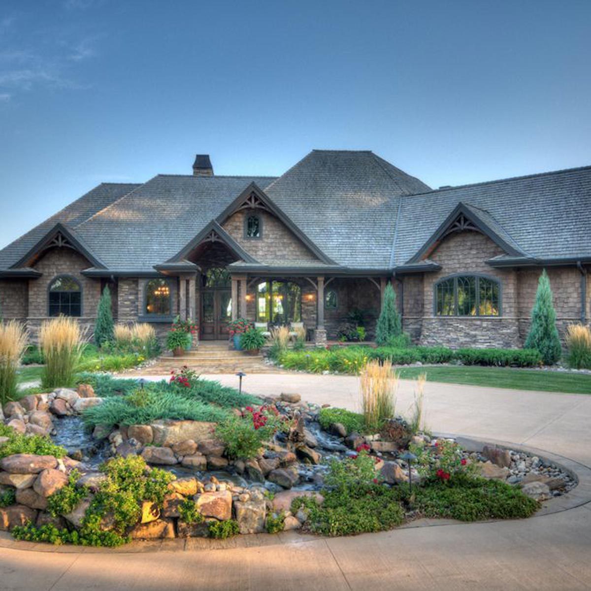 A large, rustic home with a stone exterior stands at the end of a landscaped driveway, surrounded by vibrant plants and a small decorative pond.