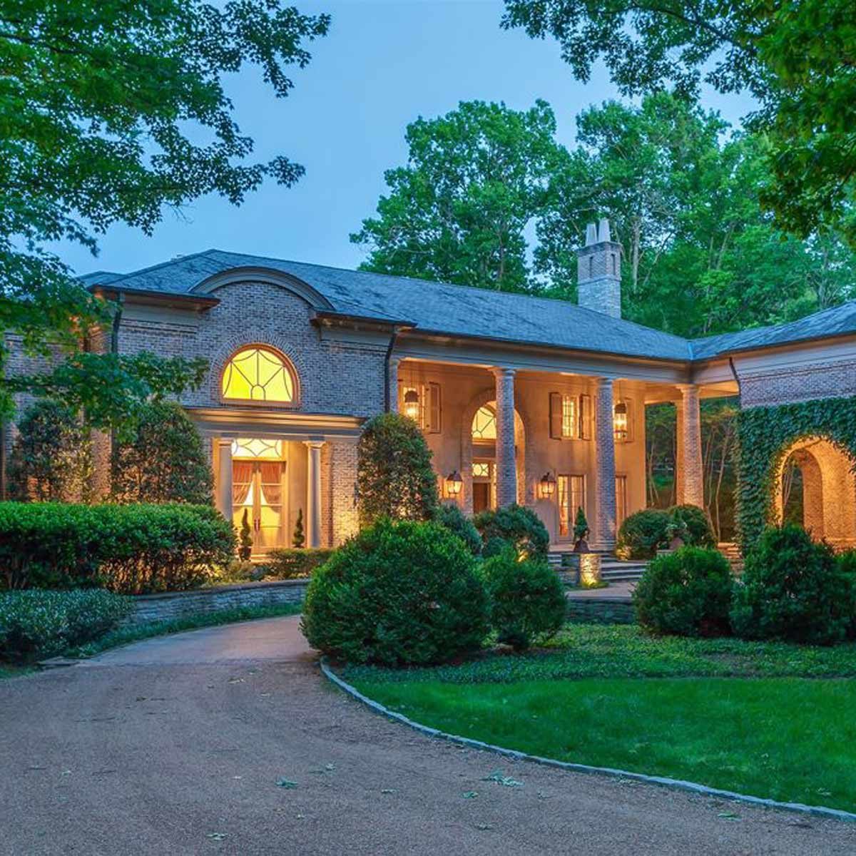 A large, elegant brick house with glowing windows, surrounded by well-manicured gardens and trees, is illuminated in the evening twilight.