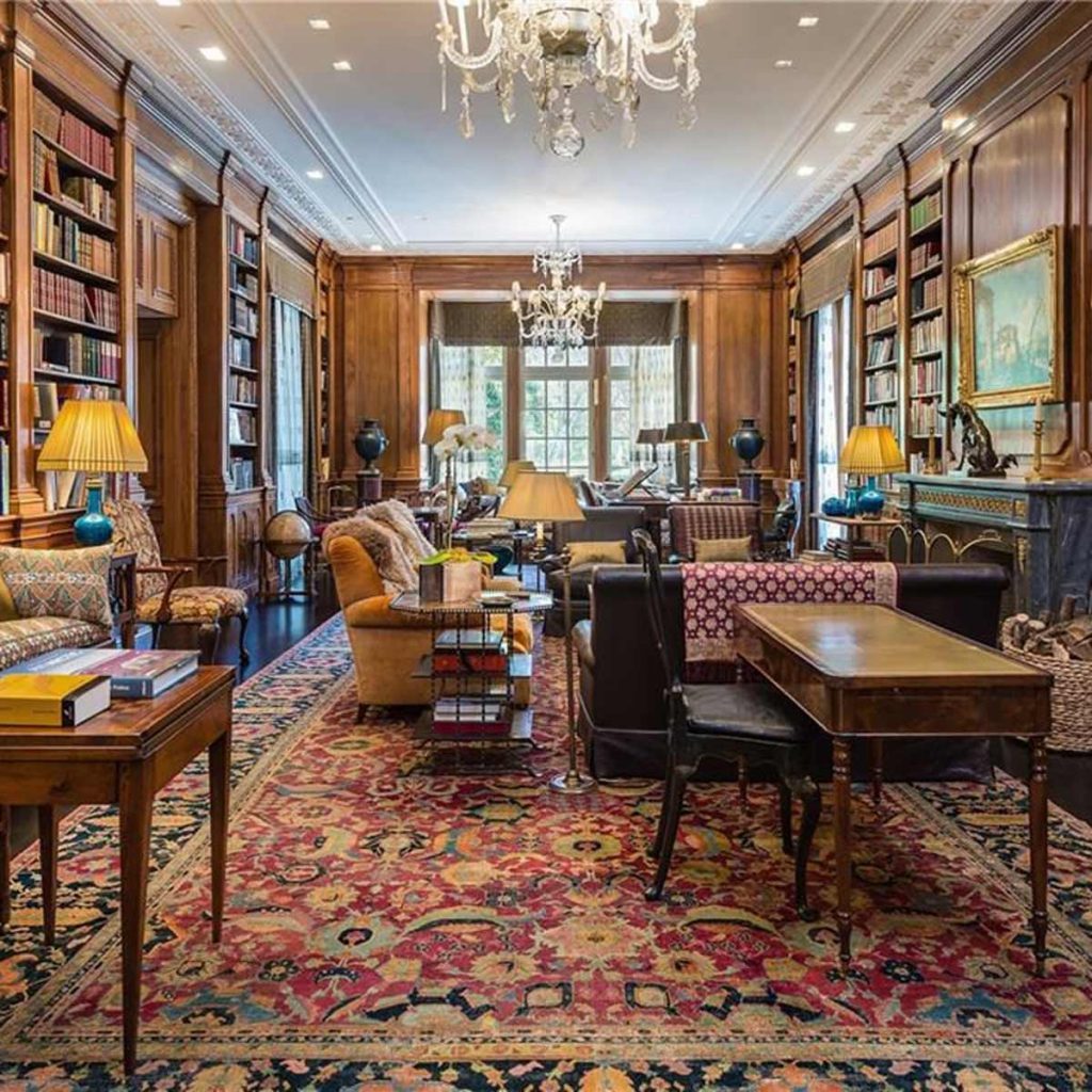 A richly furnished room features bookshelves, elegant lamps, and plush seating, creating a warm, inviting space with decorative rugs and a chandelier overhead.