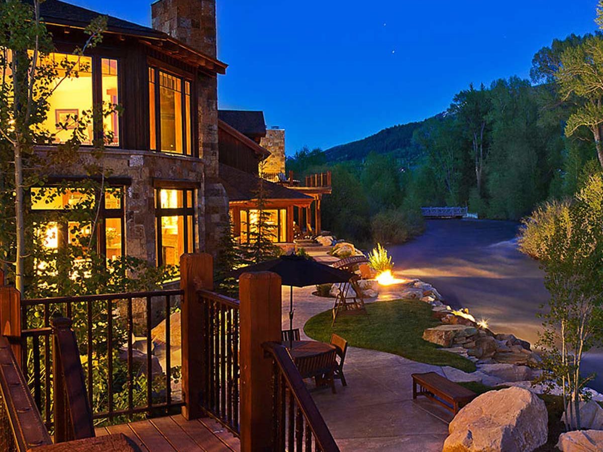 A modern riverside cabin glows warmly at dusk, with a stone exterior and wooden deck overlooking a winding waterway, surrounded by trees and landscaped stones.