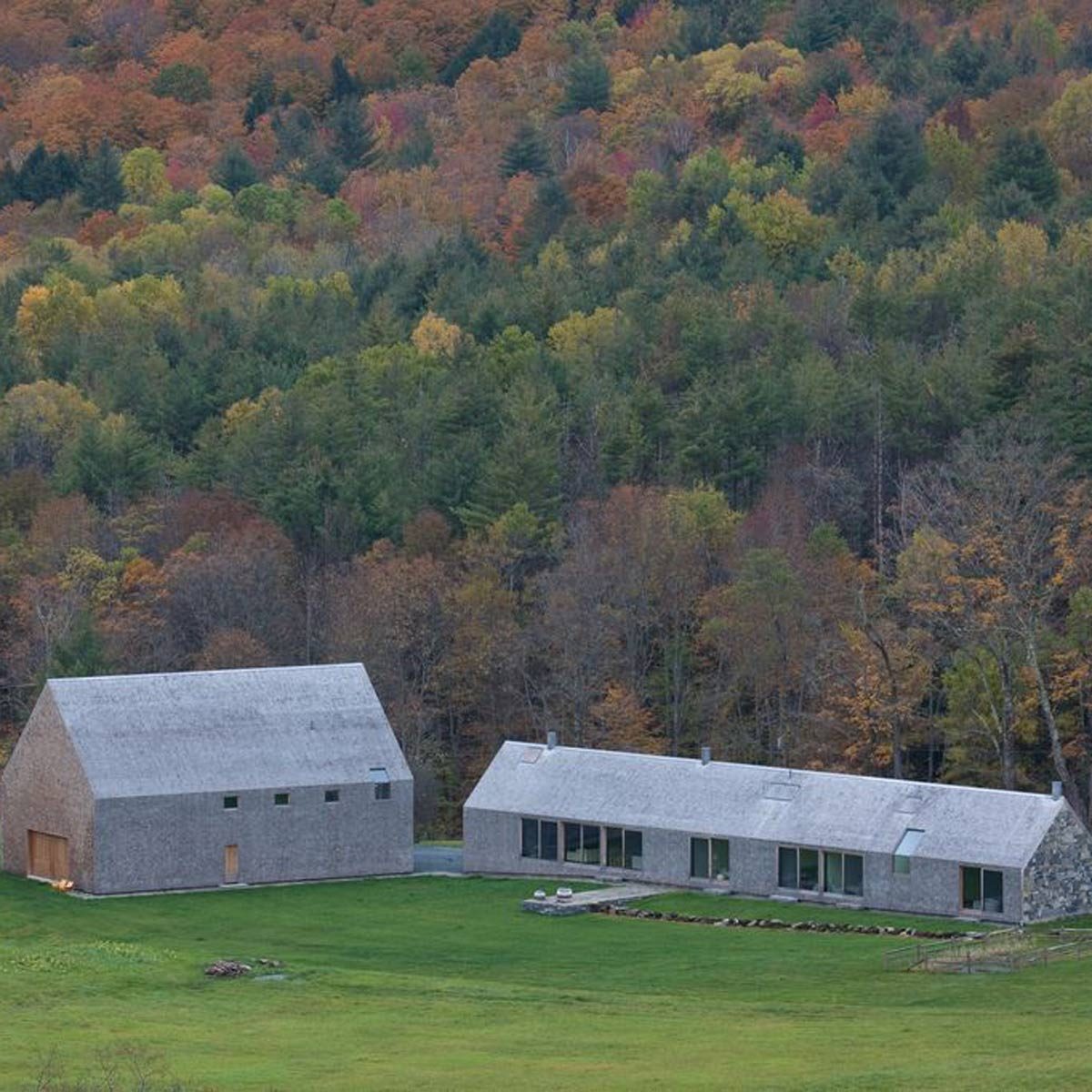 Two modern buildings with muted gray exteriors sit on a grassy area, surrounded by a forest displaying vibrant autumn colors.