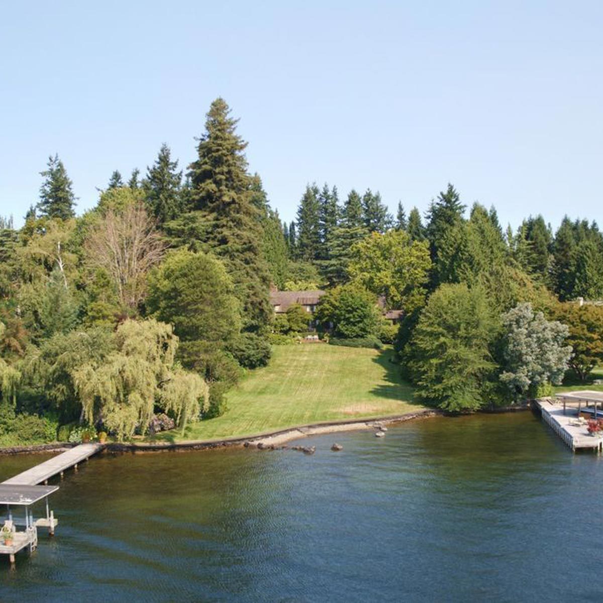 A house sits on a grassy shore beside a tranquil lake, surrounded by various trees and a wooden dock extending into the water.