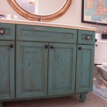 A turquoise bathroom cabinet with ornate knobs sits under a marble countertop, reflecting in a circular mirror, near a white toilet and a wall map.