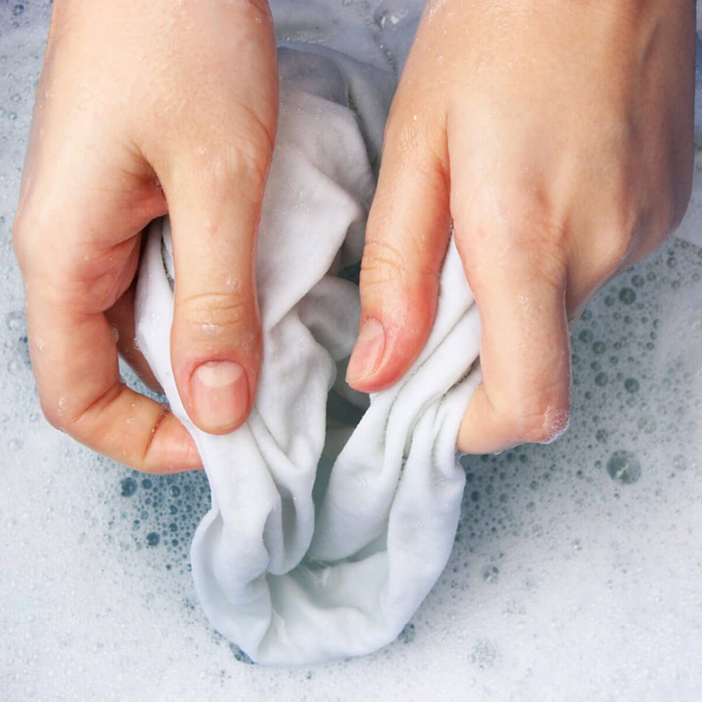 Hands squeeze a wet, white cloth, creating bubbles in soapy water. The action emphasizes the effort of cleaning within a basin filled with foam.