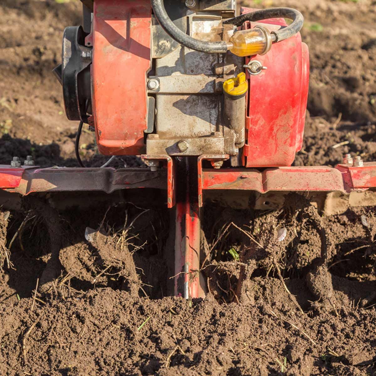 A red tiller is digging into rich, dark soil, breaking it apart and turning it over in a garden or farming context.