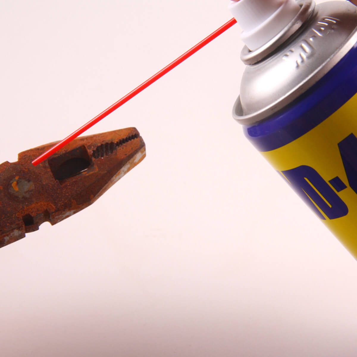A rusted pair of pliers holds a red straw while a spray can dispenses a liquid, set against a neutral background.