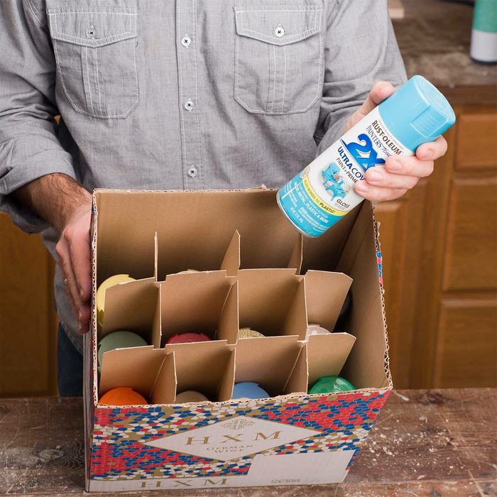 A person holds a spray can above a box containing colorful objects, organized in cardboard dividers, on a wooden surface in a workshop setting.