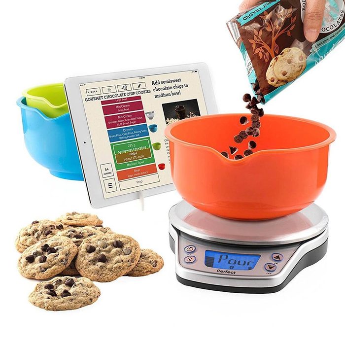 A person pours chocolate chips from a bag into an orange mixing bowl on a scale, beside a tablet displaying a cookie recipe and cookies.