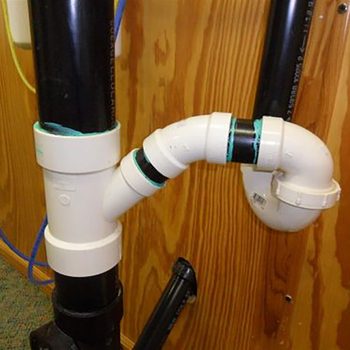 A curved white pipe connects to a black vertical pipe, both attached to a wooden wall, indicating plumbing infrastructure in a utility area.