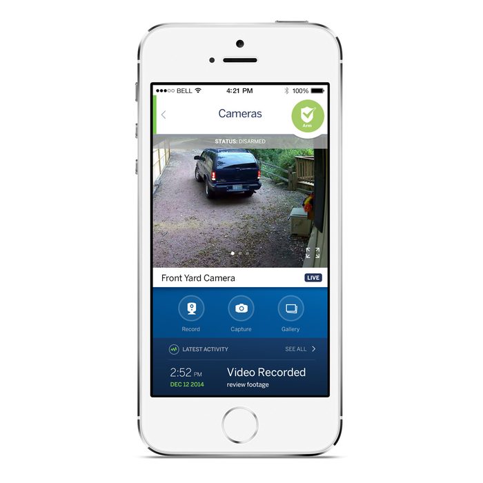 A smartphone displays a live camera feed showing a black vehicle backing out of a driveway surrounded by greenery and wooden structures.