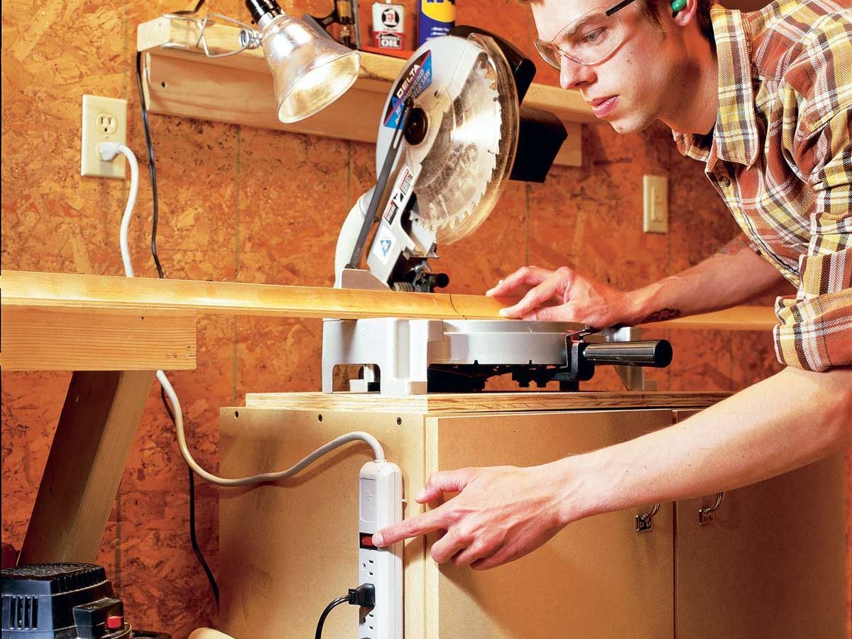 A man, wearing safety glasses, operates a miter saw on a workbench while pressing a power strip button in a workshop with wooden walls.