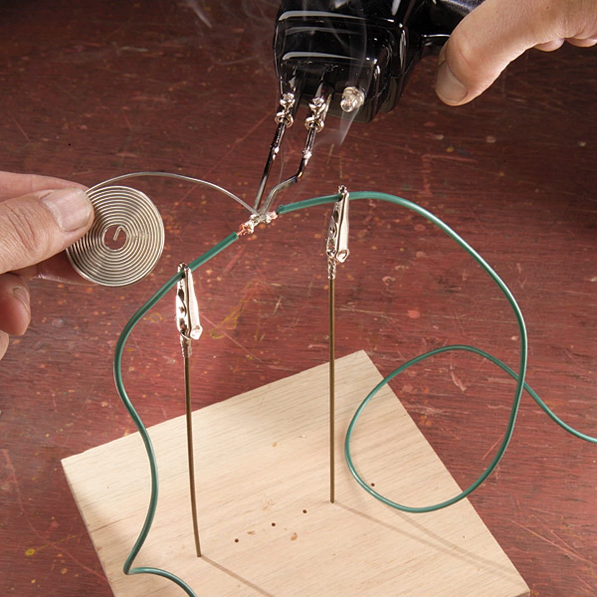 A person manipulates a metal wire using soldering tools, attaching it to a wooden base with two metal rods, set against a textured wooden surface.
