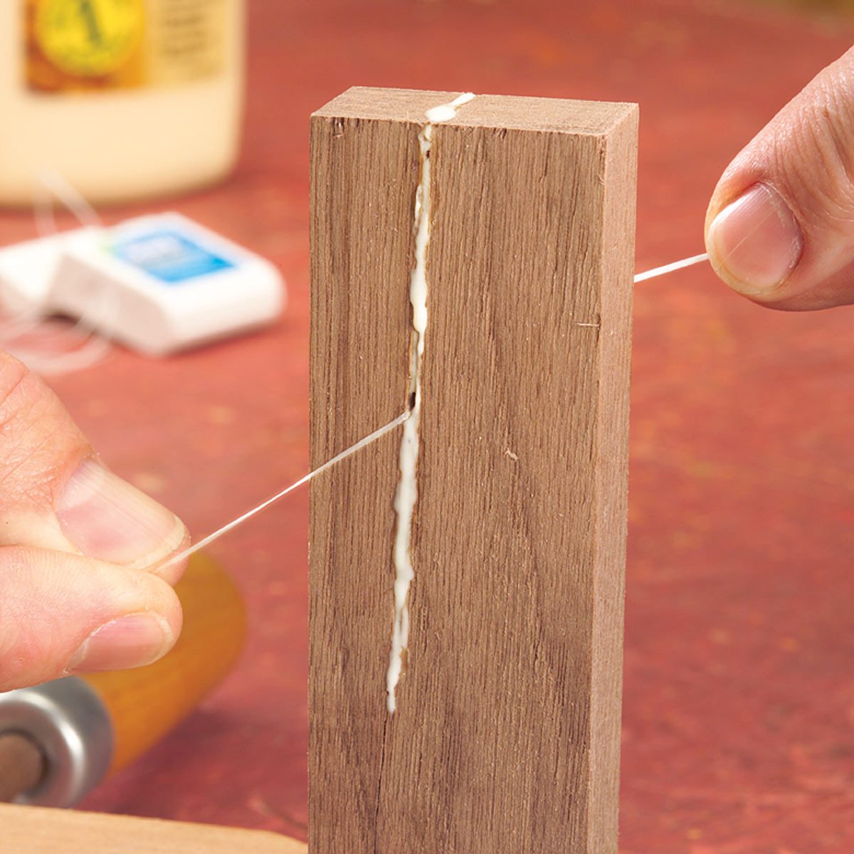 A person uses string to pull tight against a wooden piece with visible glue in a crack, amid a workshop setting with tools nearby.