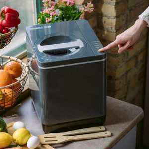 A person presses a button on a countertop food recycler, surrounded by fresh vegetables and fruits in bowls near a window with flowers.