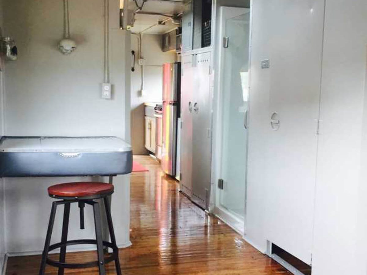 A gray sink on a stool sits in a narrow hallway leading to a kitchen, with wooden floors and bright, natural light illuminating the space.