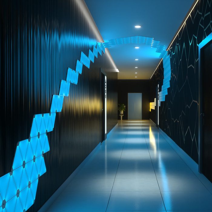 A corridor features glowing blue geometric wall panels, illuminated by ceiling lights, with doors, a plant, and a sleek modern design enhancing its ambiance.