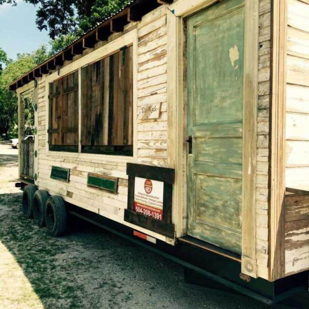 A weathered mobile home with a green door stands on wheels, partially surrounded by grass and trees, showcasing an aged appearance with wooden planks and closed windows.
