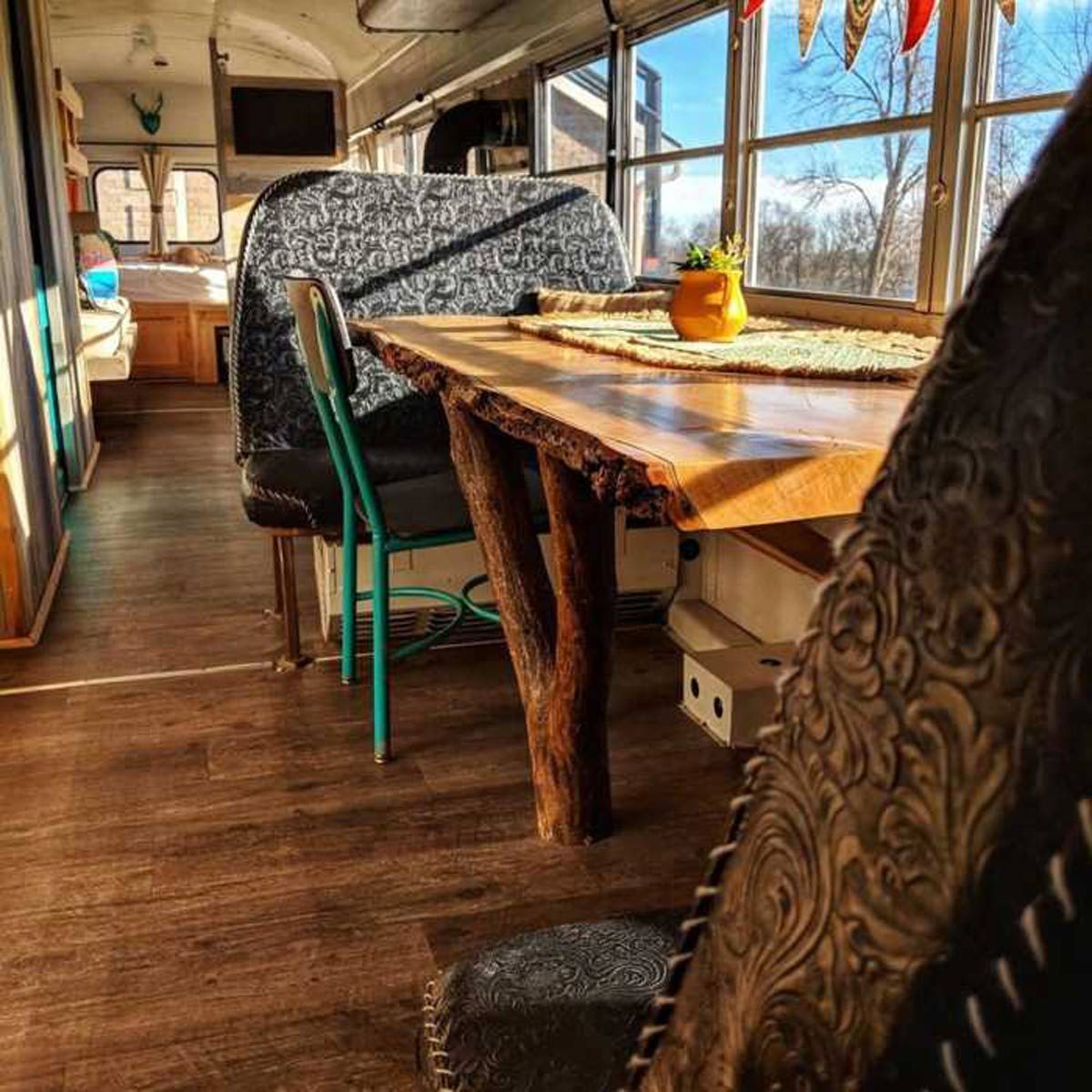 A wooden table with a unique base sits in a renovated space, surrounded by cozy seating and illuminated by natural light from large windows.