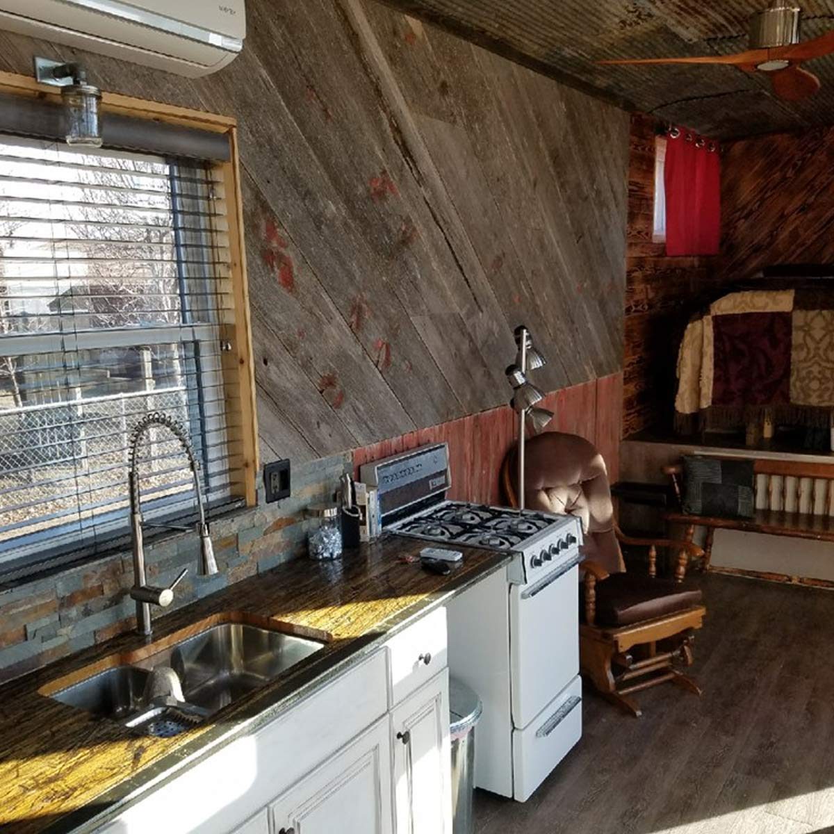 A kitchen with a sink and stove is set against rustic walls, featuring a rocking chair and a window with blinds that lets in light.