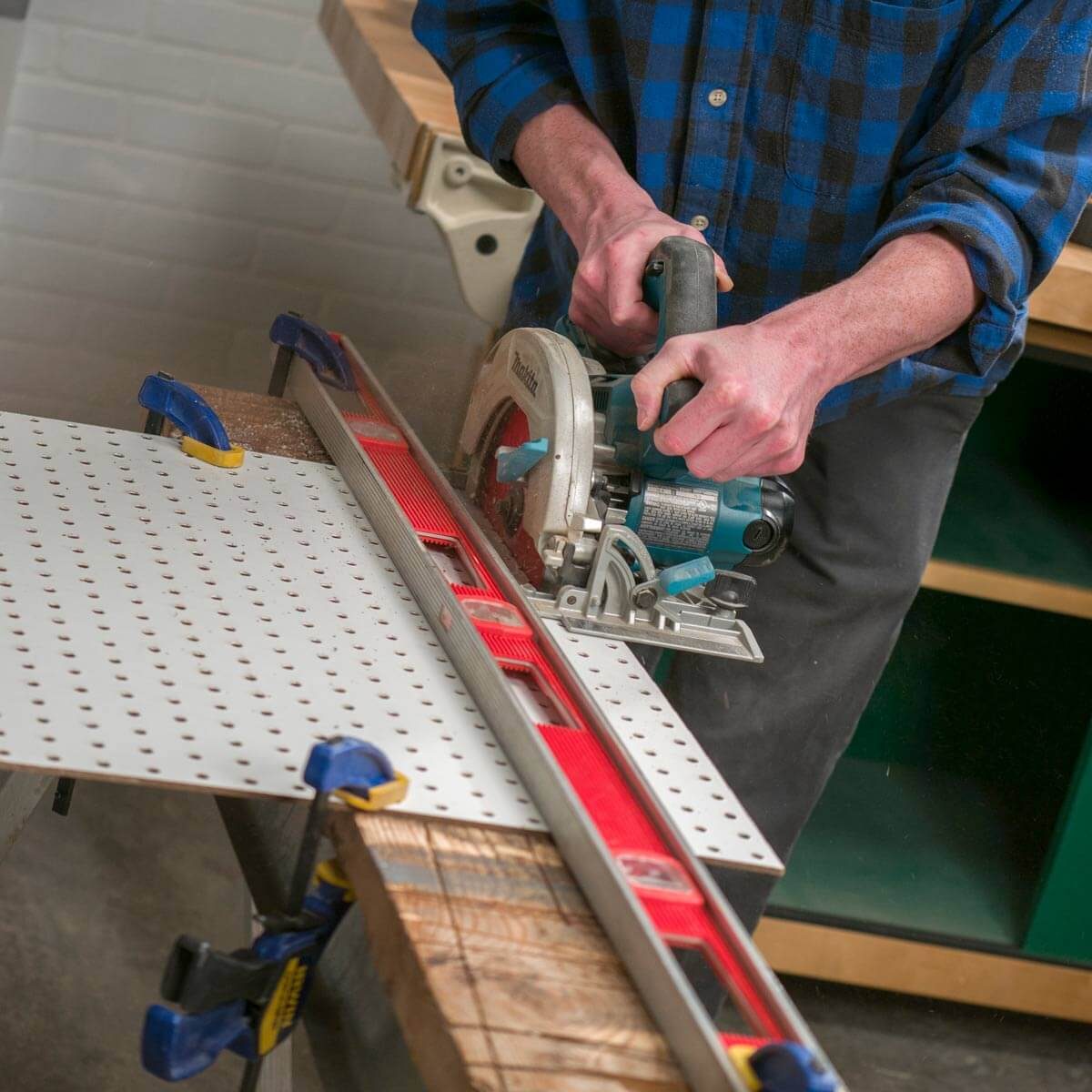 A person uses a circular saw to cut a perforated sheet secured on a workbench, with wood and tools nearby in a workshop.