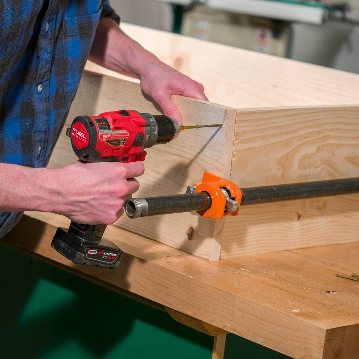 A person uses a power drill to insert a screw into a wooden board, supported by a clamp on a workbench in a workshop setting.
