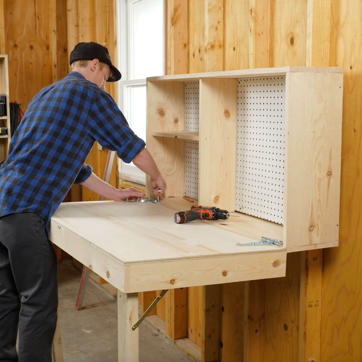 A person installs hardware on a workbench in a wooden workshop, with shelves above and tools scattered nearby, including a drill and screws.