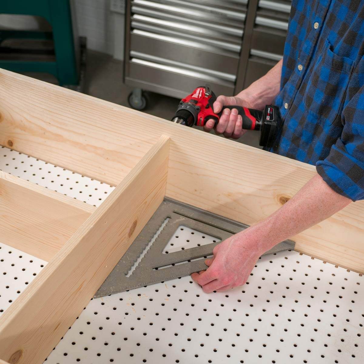 A person uses a power drill while securing a wooden frame with a square on a pegboard surface in a workshop setting.